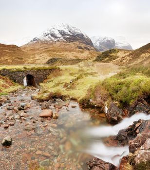 Stream and stony bridge  in spring Higland mountains in Scotland. Snowy mountains in heavy clouds. Dry grass and heather bushes on stream banks. 