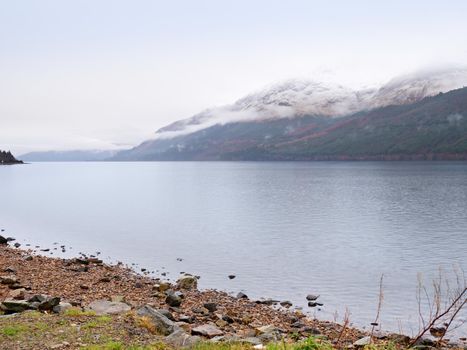 Mountain lake before misty sunset in Higland in Scotland. Snowy cone of mountain above mirroring water level. Dry grass and heather bushes on banks