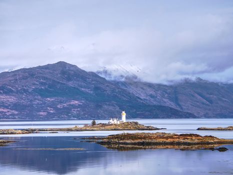 Iasle Ornsay Lighthouse built on a small islet  located on the ferry route. Low level of smooth water. Snowy mountain peaks hidden in cloudy background.  