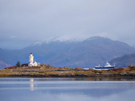 Lighthouse on Isle of Ornsay.Trade ship at rocky island, mountains in background.  Isle of Skye, Scotland, United Kingdom. 