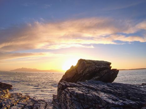 The stony beach and cliff of rocky bay. Blue tones of February sunset, pink horizon.  Dark slipery boulders with deep cracks.