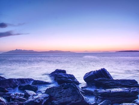 The stony beach and cliff of rocky bay. Blue tones of February sunset, pink horizon.  Dark slipery boulders with deep cracks.