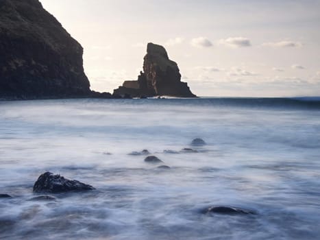 Sharp silhouette of a cliff against the background of sunset. Sea waves break on the rocks. Talisker bay, Scotland. 
