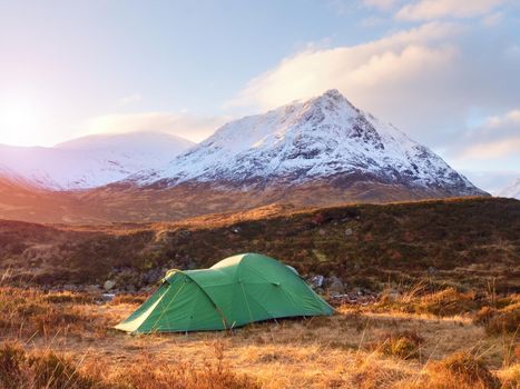 Green touristic tent on meadow at river below snowy cone of mountain Stob Dearg 1021 metres high. Higland in Scotland an marvelous sunny winter day. Dry grass and heather bushes. Hikers tent 