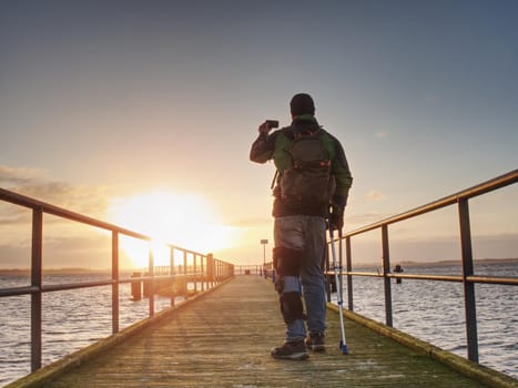 Man with phone in hand takes photo of sunrise on ferry port mole.  Hurt man with crutches.