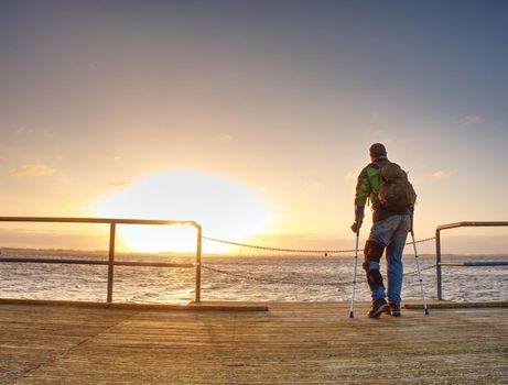 Traveler with backpack walk dificult with medicine poles.  Young tourist on bridge  near sea at the sunset