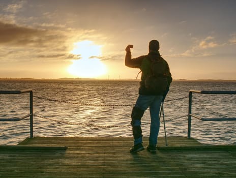 Man with phone in hand takes photo of sunrise on ferry port mole.  Hurt man with crutches.