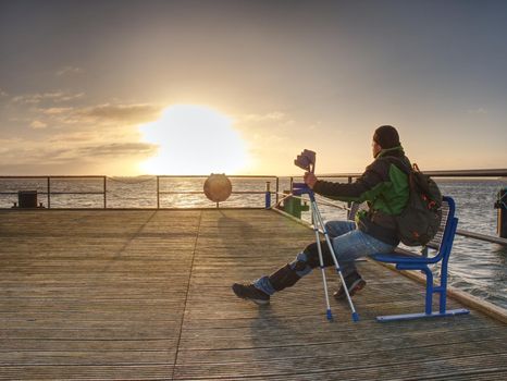 Silhouette of man with backpack on the wooden pier on the calm sea bay with sun at horizon. Beautiful golden sunset. 