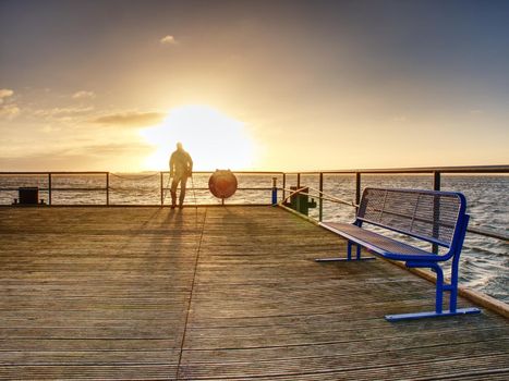 Man hiker with backpack stand alone and watching romantic colorful sunrise above wooden sea bridge