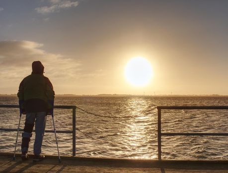 Man hiker with backpack stand alone and watching romantic colorful sunrise above wooden sea bridge