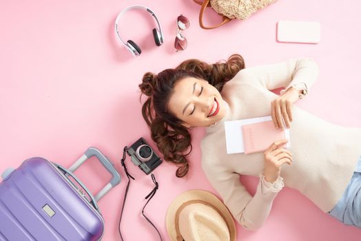 Asian woman holding her passport and lying surround her travel luggage on pink floor. Top view