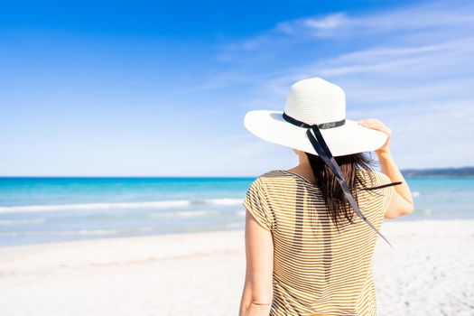 Tropical sea travel ADV banner style: unrecognizable woman holding white large hat with a hand looking to the blue ocean water standing on white silked sand under blue sky. Escape from everyday life