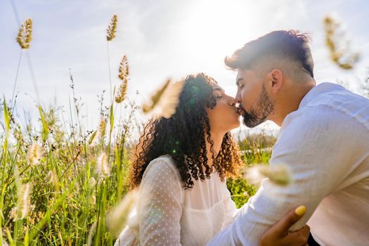 Romance scene of mixed-race couple in love kissing in backlight effect among flowers and high grass with flare and reflection sun effect at sunset or sunrise. Bearded macho young man flirting