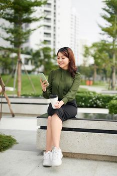 Beautiful business woman in casual green outfit sitting in park, holding coffee and talking on a phone. Daylight, outdoors.