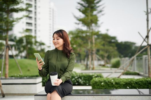 Happy young woman with a disposable coffee cup sitting on the park bench and using her smartphone