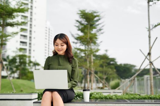 Portrait of busy sales woman sitting at business park and using her laptop while writing text on mobile. Businesswoman working online.