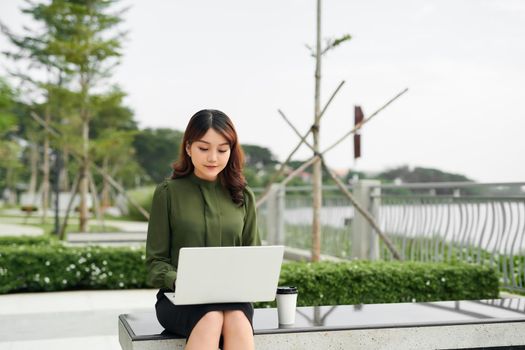 Portrait of busy sales woman sitting at business park and using her laptop while writing text on mobile. Businesswoman working online.