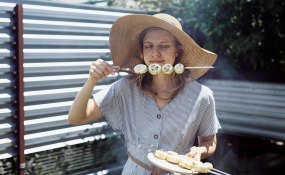 Young woman in summer hat and dress grilling meat and vegetables outdoors in the backyard. Young woman in summer hat smelling grilled vegetebles outdoors