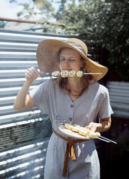 Young woman in summer hat and dress grilling meat and vegetables outdoors in the backyard. Young woman in summer hat smelling grilled vegetebles outdoors