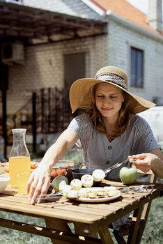 Backyard barbecue. Young woman in summer hat sitting at the table, eating grilled vegetebles outdoors