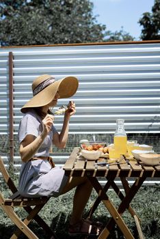 Backyard barbecue. Young woman in summer hat sitting at the table, eating grilled vegetebles outdoors