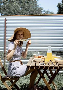 Backyard barbecue. Young woman in summer hat sitting at the table, eating grilled vegetebles outdoors