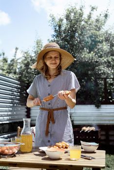 Young woman in summer hat and dress grilling meat and vegetables outdoors in the backyard.