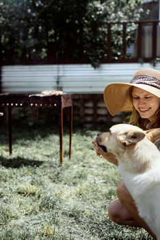 Young woman in summer hat grilling meat outdoors in the backyard, sitting with her dog, giving pet a snack