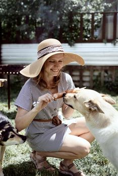 Young woman in summer hat grilling meat outdoors in the backyard, sitting with her dog, giving pet a snack