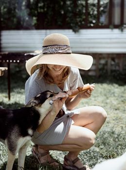 Young woman in summer hat grilling meat outdoors in the backyard, sitting with her dog, giving pet a snack