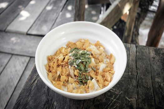 healthy breakfast fruit bowl with banana, corn flakes, pumpkin seeds and coconut milk on rustic wood table outdoors