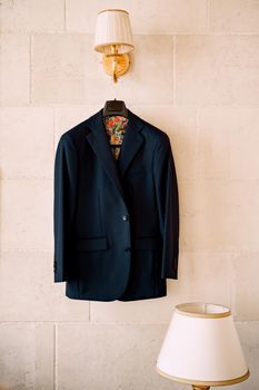 A blue jacket with colorful lining on a black hanger hangs from a wall lamp in a room with a table lamp. High quality photo