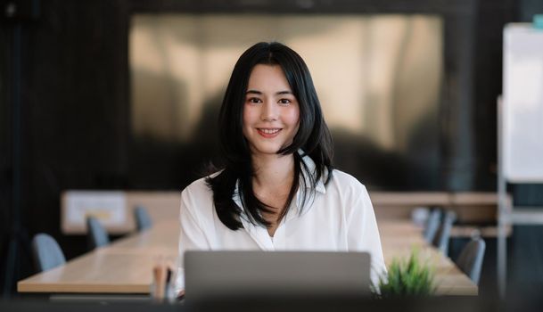 Smiling young asian businesswoman sitting on her workplace in the office. Young woman working at laptop in the office.