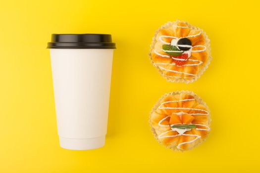 Creative flat lay with White cardboard coffee cup and two mini cakes with pieces of fruits and cream on yellow background. Concept of fast carbs and unhealthy snacks