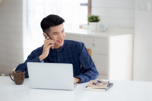 Young asian man talking phone and work from home with laptop computer, freelance using notebook to internet online and speak on smartphone, domestic life, business and communication concept.