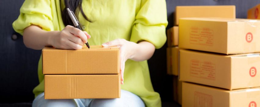Closeup hand of young asian woman writing address on parcel box at home for delivery order to customer, shipping and logistic, merchant online and seller, business owner or SME, online shopping.