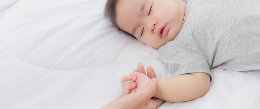 Young asian mother touch hand of little baby girl with tender on bed in the bedroom, mom love newborn and care, woman with expression with child together, parent and daughter, family concept.