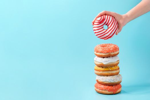 Stack of assorted donuts and one donut in female hand on blue background. Many colorful glazed doughnut with sprinkles stacked with copy space