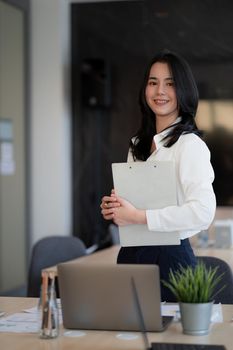 Portrait of successful young Asian businesswoman standing at office, colleagues in background.