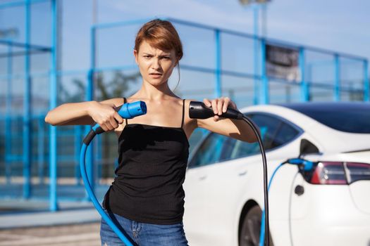 Beautiful young girl next to an electric car. Confused with two charging cables