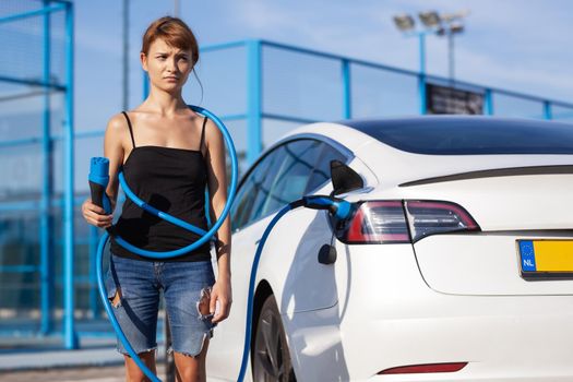 Beautiful young girl next to an electric car. Clumsy wrapped and confused with a charging cable around her body.