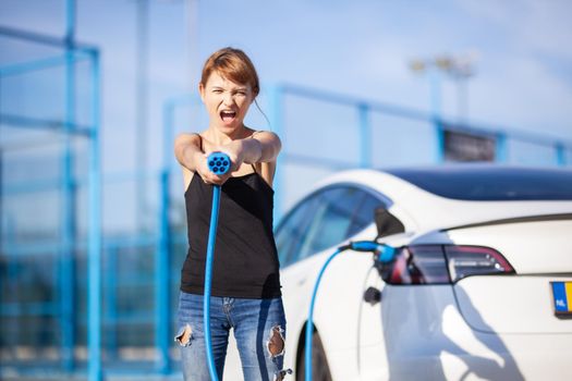 Beautiful young girl next to an electric car. Posing and holding a charging cable.