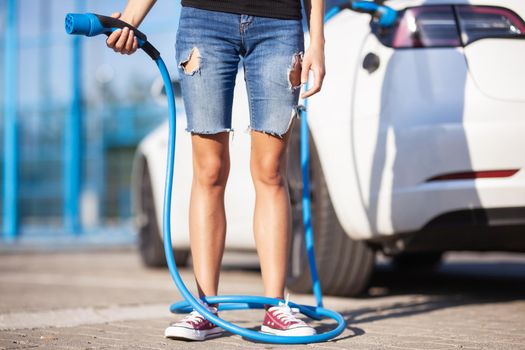 Young girl next to an electric car. Clumsy wrapped with a charging cable around her legs.