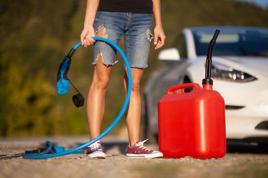 Girl standing next to an electric car. Holding charging cable and gassoline canister.