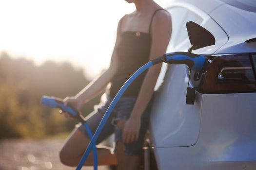 Beautiful young girl next to an electric car. Holding a charging cable. Sunset backlight.