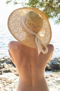 Rear view of topless beautiful woman wearing nothing but straw sun hat realaxing on wild coast of Adriatic sea on a beach in shade of pine tree. FKK sunbathing lifestyle concept