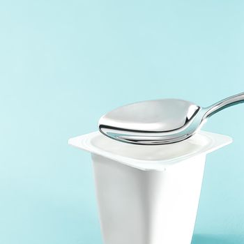 Yogurt cup and silver spoon on mint background, white plastic container with yoghurt cream, fresh dairy product for healthy diet and nutrition balance.