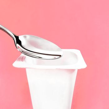 Yogurt cup and silver spoon on pink background, white plastic container with yoghurt cream, fresh dairy product for healthy diet and nutrition balance.