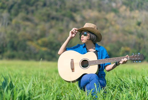 Young asian women short hair wear hat and sunglasses sit playing guitar in grass field countryside Thailand