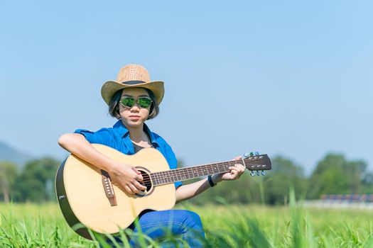 Young asian women short hair wear hat and sunglasses sit playing guitar in grass field countryside Thailand